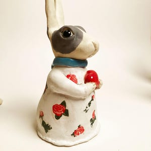 Może przedstawiać: Ceramiczna figurka królika z długimi uszami, szarą plamką nad jednym okiem i niebieską obrożą. Królik ma na sobie białą sukienkę ozdobioną małymi wzorami czerwonych róż i trzyma w przednich łapkach małe, gładkie, błyszczące czerwone serce.