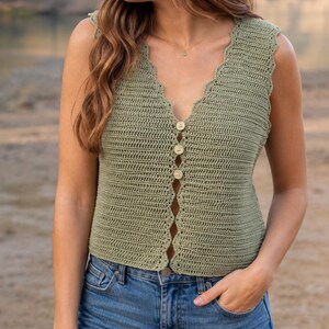 May include: A sleeveless, sage green crochet top with a scalloped neckline and button-up front. The top is paired with blue jeans. The crochet stitch creates a textured appearance. The buttons are a light beige color.