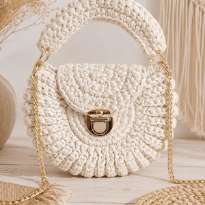 May include: A round, off-white crochet handbag featuring a thick, textured woven handle and a gold-tone metal push-lock closure. The bag includes a detachable gold-tone metal chain shoulder strap attached to the sides with circular metal rings.