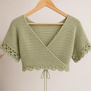 May include: A sage green crochet wrap top with short sleeves and a scalloped edge. The top is hanging on a wooden hanger. The wrap design features a V-neck and ties at the waist. The crochet stitch creates a textured appearance.