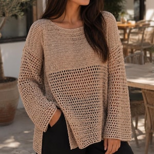 Könnte beinhalten: Ein beigefarbener, langärmeliger Häkelpullover mit einem lockeren, offenen Strickmuster an Körper und Ärmeln, einem weiten Rundhalsausschnitt und Seitenschlitzen am Saum, kombiniert mit einer schlichten schwarzen Hose.