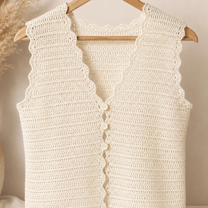 May include: Cream-colored, sleeveless crochet vest with a scalloped edge and a V-neck. The vest has a button-up front with four matching buttons. The garment is hanging on a wooden hanger.