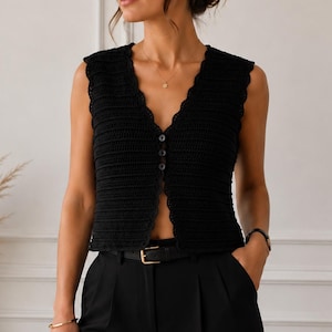 Peut inclure: Un gilet sans manches en crochet noir doté d'une encolure en V avec un bord festonné et trois boutons noirs assortis sur le devant. Le vêtement présente un motif en maille côtelée texturée sur toute la surface et est porté avec un pantalon noir.