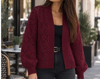 Könnte beinhalten: Eine tiefburgunderrote, offene Strickjacke mit strukturiertem Rautenmuster, langen Ballonärmeln mit gerippten Bündchen und einem breiten Rippstricksaum entlang der Vorderseite. Das Kleidungsstück wird über einem schwarzen Oberteil und einer dunkelgrauen Jeans getragen.