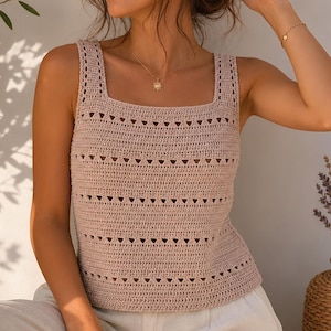 May include: A light tan crochet tank top with a square neckline and openwork detailing. The top has a textured appearance and is paired with light-coloured trousers. A gold necklace with a sun pendant and gold earrings are visible.