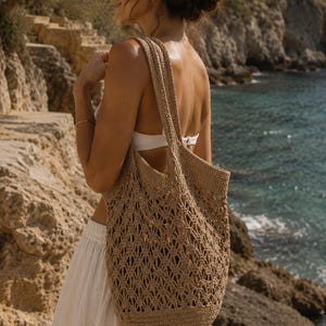 Puede incluir: Un bolso tote de ganchillo beige que presenta un intrincado patrón de diamantes calados en todo el cuerpo, una base circular sólida y dos correas largas para el hombro. El bolso está construido con un material de hilo texturizado de color tostado.