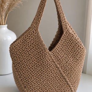 Puede incluir: Un bolso de ganchillo color canela con un patrón tejido texturizado y un asa integrada única. El bolso presenta un detalle de costura diagonal distintivo en la parte frontal, creando una forma estructurada y asimétrica con una abertura amplia.