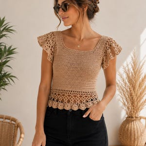 May include: A tan crochet top with a square neckline and short, ruffled sleeves. The top has a scalloped hem and is paired with black jeans. The image is taken in natural light, showcasing the texture of the crochet.