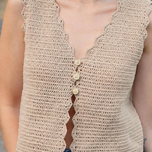 May include: A beige, sleeveless crochet top with a scalloped neckline and hem. The top features a V-neck and is fastened with three wooden buttons. The crochet stitch creates a textured appearance, and the garment is suitable for warm weather.