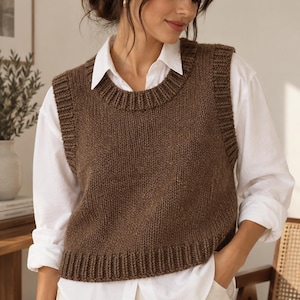 Könnte beinhalten: Eine braune, ärmellose Strickweste mit einem dicken, gerippten Rundhalsausschnitt, gerippten Armausschnitten und einem gerippten Saum. Das Kleidungsstück wird über einem weißen, langärmeligen Hemd mit Knopfleiste und einer hellen Hose getragen.