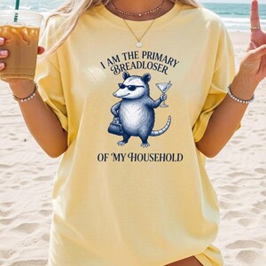 May include: A pale yellow t-shirt featuring a graphic of an opossum wearing sunglasses, holding a martini glass and a small handbag. The text above and below the opossum reads, I AM THE PRIMARY BREADLOSER OF MY HOUSEHOLD in dark blue lettering.