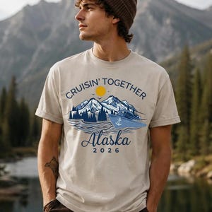 May include: A beige short-sleeve t-shirt featuring a graphic print of mountains, a cruise ship, and a yellow sun. The text reads "CRUISIN' TOGETHER" in an arc above the mountains, with "Alaska" and "2026" printed in blue below the ship.