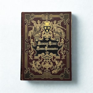 Puede incluir: Un libro antiguo con una cubierta burdeos y detalles dorados ornamentados. La portada presenta el texto "MODERNE KUNST in Meister-Holzschnitten" en letras doradas, junto con ilustraciones de figuras aladas y adornos decorativos.