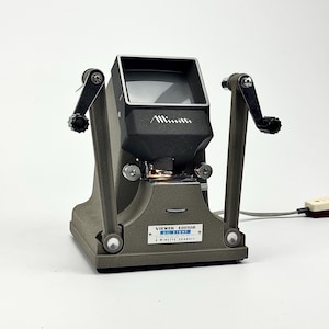 Può includere: Un vecchio visualizzatore di film Minette Viewer Editor in grigio e nero. Il dispositivo ha uno schermo di visualizzazione, due manovelle e un'etichetta che recita "Viewer Editor Swift Light A Minette Product."