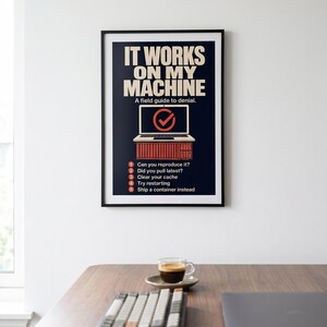 May include: A framed poster with the text "IT WORKS ON MY MACHINE" and "A field guide to denial." The poster includes a laptop graphic and a list of troubleshooting steps. A cup of coffee, keyboard, and laptop are on a wooden desk.
