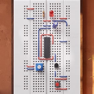 May include: A white breadboard mounted in a dark wood frame featuring an integrated circuit, a red LED, a blue capacitor, a resistor, a blue potentiometer, a black transistor, and a red push button connected by red and blue jumper wires.
