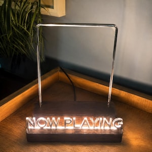Könnte beinhalten: Ein Holzsockel mit den Worten "NOW PLAYING" in Weiß beleuchtet. Ein klarer, rechteckiger Rahmen erhebt sich von der Basis, mit einem hellen Licht an der Oberseite. Die Lampe steht auf einer Holzoberfläche.