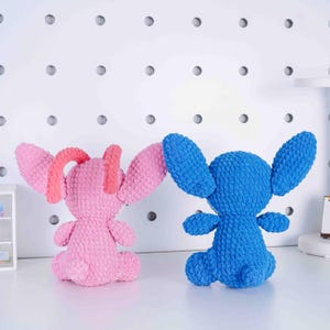 May include: Two handmade crochet plush toys. One is pink with coral accents, the other is blue. Both have large ears, small arms, and are seated. The toys are on a white surface with a white pegboard background.
