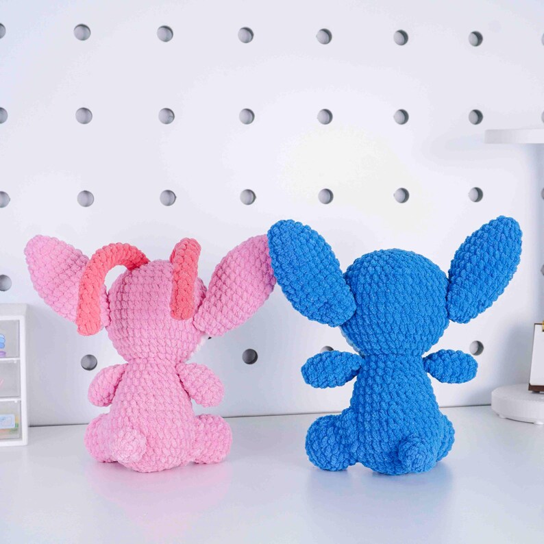 May include: Two handmade crochet plush toys. One is pink with coral accents, the other is blue. Both have large ears, small arms, and are seated. The toys are on a white surface with a white pegboard background.