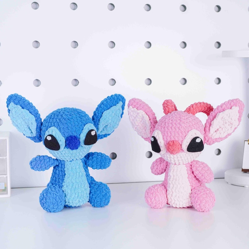 May include: Two handmade crochet plush toys, one blue and one pink, resembling cartoon characters. The blue plush has large ears and a darker blue snout. The pink plush has similar features with a lighter pink color scheme. Both have black eyes and are sitting upright.