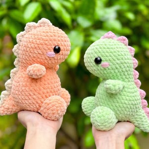 Könnte beinhalten: Zwei handgefertigte Dinosaurier-Plüschtiere aus Häkelarbeit. Eines ist pfirsichfarben, das andere grün. Beide haben schwarze Augen, rosa Akzente und werden vor einem grünen, belaubten Hintergrund gehalten. Diese weichen Spielzeuge sind perfekt für Kinder.