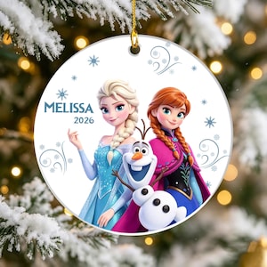 May include: A round white ceramic ornament featuring Elsa, Anna, and Olaf from Frozen. The text MELISSA and 2026 is printed on the left side. The design includes blue snowflake patterns and decorative swirls, suspended by a gold string.