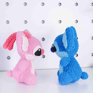 May include: Two crocheted plush toys, one pink and one blue, are positioned facing each other. The pink toy has long ears with pink accents, black eyes, and a pink nose. The blue toy has large ears, black eyes, and a blue nose. Both toys are made of a soft, textured yarn.