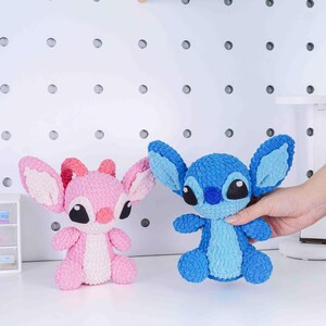 May include: Two plush toys, one pink and one blue, resembling cartoon characters. The pink toy has a lighter pink inner ear and a darker pink antenna. The blue toy has large ears and is held by a hand. Both toys have black eyes.