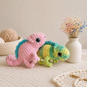 Pode incluir: Dois camaleões de crochê feitos à mão. Um é rosa com uma espinha dorsal azul, o outro é verde e amarelo com uma espinha dorsal azul. Ambos têm olhos grandes e pretos. Os camaleões estão em uma superfície branca.
