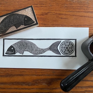 Puede incluir: Un grabado en linóleo en blanco y negro de un pez con un diseño de nudo celta. La impresión está sobre papel blanco, con un bloque de madera y un rodillo de tinta negro en la imagen. El pez está en un marco rectangular.
