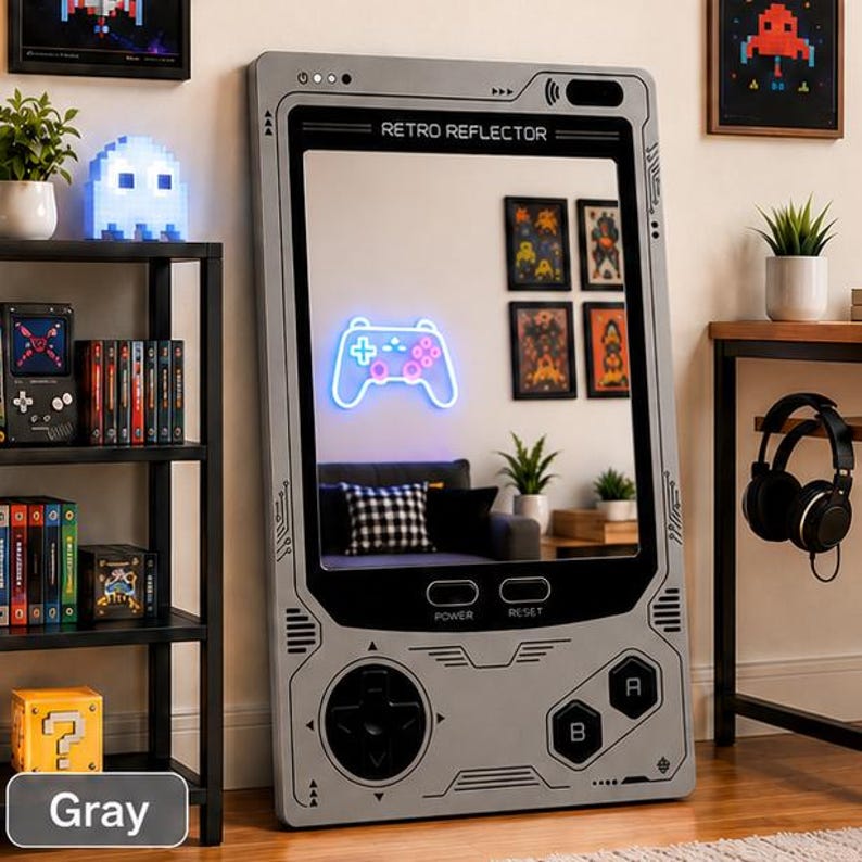 May include: A large rectangular floor mirror designed to resemble a vintage handheld gaming console. The gray frame features printed buttons, a directional pad, and the text RETRO REFLECTOR, POWER, and RESET. The mirror reflects a room interior.