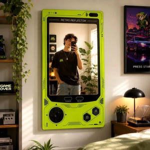 May include: A rectangular wall mirror designed to resemble a handheld gaming console in bright lime green. The frame features black directional buttons, A and B buttons, and power and reset labels. The top of the frame displays the text RETRO REFLECTOR.