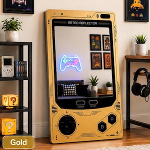 May include: A rectangular gold-colored mirror designed to resemble a handheld gaming console. The frame features black buttons, a directional pad, and the text RETRO REFLECTOR. The mirror surface reflects a room with a glowing neon game controller.