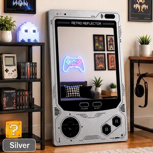 May include: A large, rectangular, silver-colored floor mirror designed to resemble a vintage handheld gaming console. The frame features a directional pad, A and B buttons, and power and reset labels. The screen area displays the text RETRO REFLECTOR.