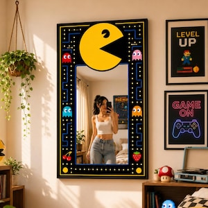 May include: A rectangular wall mirror featuring a black frame decorated with a yellow Pac-Man character, colorful pixelated ghosts, cherries, and strawberries. The border design mimics a classic arcade maze with yellow dots on a black background.