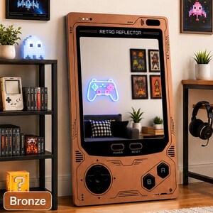 May include: A large rectangular mirror designed to resemble a handheld gaming console with a bronze-colored frame. The frame features printed buttons, a directional pad, and the text RETRO REFLECTOR, POWER, and RESET. The mirror reflects a room.
