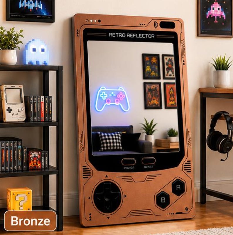 May include: A large rectangular mirror designed to resemble a handheld gaming console with a bronze-colored frame. The frame features printed buttons, a directional pad, and the text RETRO REFLECTOR, POWER, and RESET. The mirror reflects a room.
