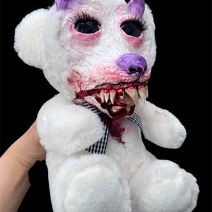 Pode incluir: Um urso de peluche branco com um rosto esculpido e texturizado, apresentando chifres roxos, longos cílios pretos e uma boca aberta revelando dentes afiados. A área do rosto e da boca tem detalhes com pintura vermelha semelhante a sangue.