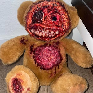 Pode incluir: Um urso de peluche castanho com um rosto modificado, tecido vermelho exposto, um olho visível e uma boca larga cheia de dentes realistas. O peito e um pé apresentam feridas texturizadas e sangrentas com detalhes de aspeto muscular.