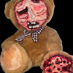 Könnte beinhalten: Ein brauner Plüschteddybär mit einem modifizierten Gesicht aus rotem und weißem, texturiertem Muskelgewebe, einem sichtbaren Auge und menschenähnlichen Zähnen. Ein schwarz-weiß kariertes Band ist um den Hals gebunden, und ein Fuß ist durch eine kreisförmige, poröse, rot und dunkel gefärbte Oberfläche ersetzt.