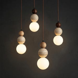 Könnte beinhalten: Vier Pendelleuchten mit modernem Design. Jede Leuchte besteht aus einer großen, runden, mattierten Glaskugel, die mit einer steinartigen Kugel, einer Holzkugel und einer dunkelbraunen Holzkugel gestapelt ist. Sie hängen an braunen Kabeln.