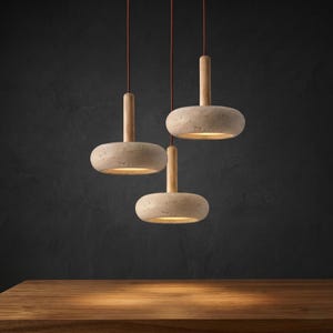 May include: Three pendant lights with stone-effect shades and wooden stems, suspended from red cords. The lights are illuminated, casting a warm glow. The background is a dark, textured wall, and the lights are above a wooden surface.