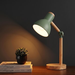 May include: A desk lamp with a sage green metal shade and a light-toned wooden base, arm, and accents. The lamp features an adjustable joint with a tightening knob and emits a warm light. A small potted plant sits on two stacked books.