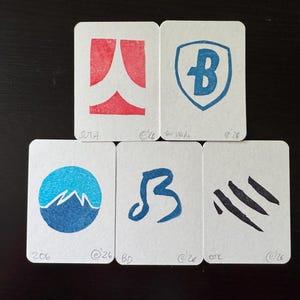 Op de afbeelding: Vijf witte rechthoekige kaarten met abstracte ontwerpen in rode en blauwe inkt. De ontwerpen omvatten een schild met een B, een berglandschap en andere geometrische vormen. Elke kaart heeft tekst.