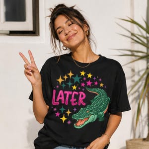 Może przedstawiać: Czarny t-shirt z zieloną grafiką aligatora i różowym napisem "LATER". Projekt zawiera kolorowe gwiazdy. Swobodny styl unisex.