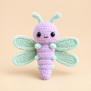 May include: A handcrafted crochet dragonfly plush toy featuring a lavender-colored body and head with mint green wings and antennae. The creature has two large black safety eyes, a small embroidered mouth, and soft pink blush on its cheeks.