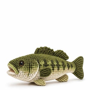 May include: A crocheted fish toy featuring a dark olive green back with a textured scale pattern, a cream-colored underbelly, and a prominent dorsal fin. The fish has a single dark brown plastic eye and an open mouth showing light interior stitching.