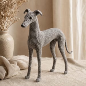 May include: A grey crochet dog figurine featuring a slender body, long legs, a pointed snout, and dark plastic eyes. The handmade textile sculpture is crafted with a tight stitch pattern, capturing the distinct silhouette of a sighthound breed.