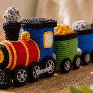 Könnte beinhalten: Ein mehrfarbiger Häkel-Spielzeugeisenbahnzug, bestehend aus einer Lokomotive mit einer grauen Rauchwolke und drei angehängten Waggons. Die Lok ist rot, blau und gelb, während die Waggons grün, blau und rot mit gelber Ladung gestaltet sind.