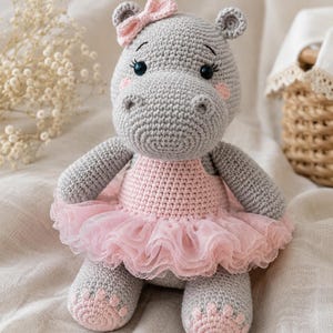 May include: A handmade gray crochet hippo wearing a pink tutu and a matching bow. The hippo has pink accents on its cheeks and feet, and black eyes. The toy is sitting on a neutral-colored surface.